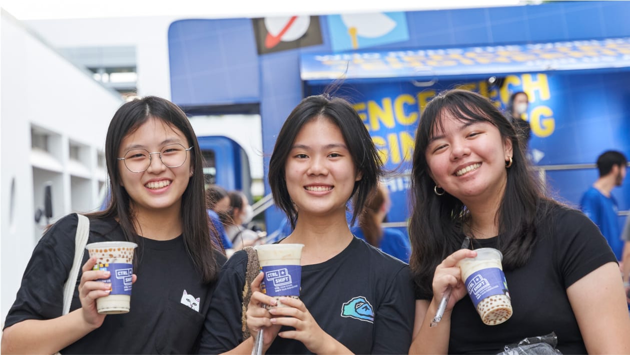 Three smiling young women in black shirts hold bubble tea cups outdoors near a blue booth.